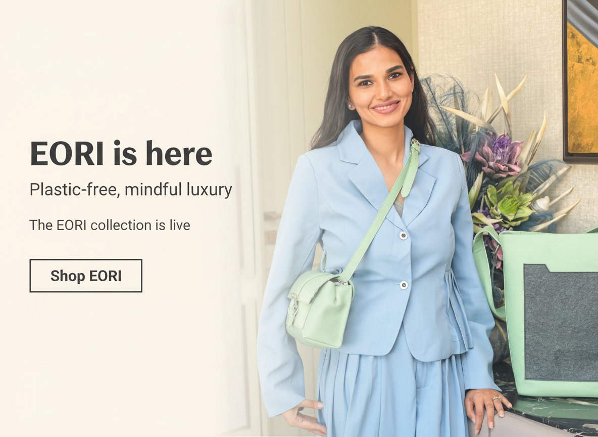 Woman in a light blue suit with a green handbag, standing indoors with plants in the background.